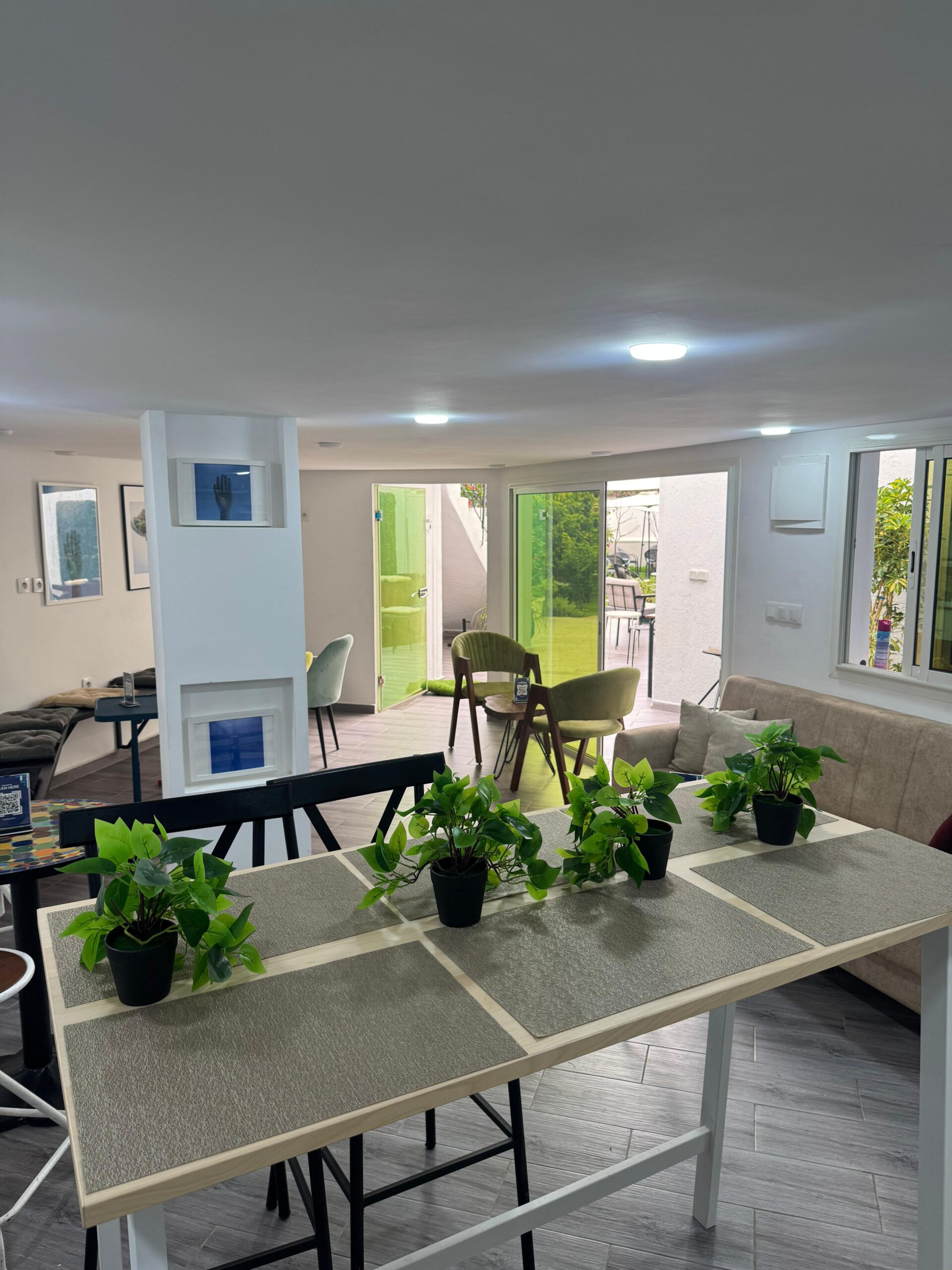 A bright and inviting lounge or break area at Al Hub Coworking Space in Rabat, featuring a long table with plants, comfortable seating, and a view to an outdoor patio with green doors.