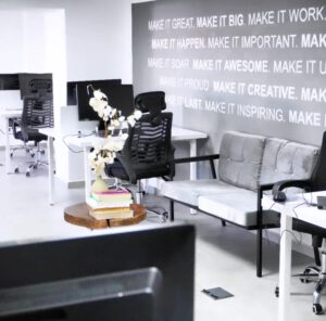 A modern office space with white desks, black ergonomic chairs, and a large motivational quote mural on a gray wall.