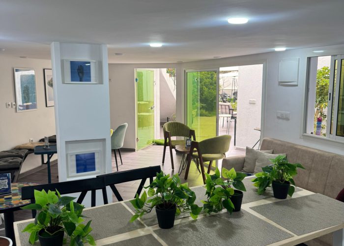 A bright and inviting lounge or break area at Al Hub Coworking Space in Rabat, featuring a long table with plants, comfortable seating, and a view to an outdoor patio with green doors.